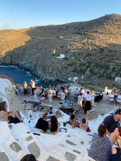 The image depicts an outdoor gathering set on multi-tiered, whitewashed stone platforms and steps, overlooking a rocky coastline and the Aegean Sea, characteristic of a location in Απολλωνία, Greece. The scene is illuminated by the late afternoon sun, casting distinct shadows.
Approximately 35 individuals, primarily adults, are present. In the mid-ground, a central area features several round and square tables with dark chairs, where guests are seated, consuming beverages and food from plates and glasses. Bottles and tableware are visible on the tables. A serving station or bar is equipped with multiple bottles, an ice bucket, and glassware. A male in a white collared shirt and black trousers, consistent with a server, is attending to guests near a table. Among standing attendees, a woman in white shorts and a dark top is captured in mid-movement, and a man plays a saxophone.
The foreground shows several individuals seated on descending whitewashed stone steps. A woman in a striped tank top and dark shorts is seated on an upper step. Further down, a man in a black t-shirt is seated beside a woman with long dark hair. These individuals are either observing the main gathering or conversing.
The background consists of arid, terraced hillsides with sparse vegetation, featuring scattered white buildings, some with blue accents, extending towards the sea. The sea itself is deep blue with whitecaps breaking against a rugged, rocky shore.