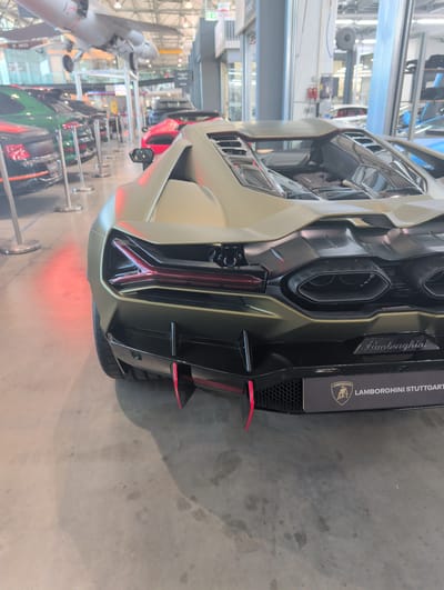 The image shows the rear of a tan-colored Lamborghini sports car. The car is parked indoors, likely in a showroom or exhibition hall, given the presence of other vehicles and display elements in the background. The car's distinctive taillights are illuminated, casting a red glow on the concrete floor. The car features prominent exhaust pipes and aerodynamic fins on the rear bumper, painted in red. A black panel above the bumper displays the Lamborghini logo and text that reads "LAMBORGHINI STUTTGART". The overall impression is one of a high-performance vehicle on display. The scene appears to be well-lit, suggesting an indoor event. The image is taken from a low angle, emphasizing the car's aggressive design.