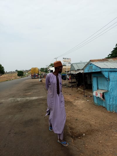 An adult male, dressed in a light purple traditional garment and a brown woven hat, stands facing right and looking forward, his body angled slightly away from the camera. He is wearing blue flip-flops. The man is positioned on the right side of a dusty road in Gadao, Nigeria. To his right, a small, vibrant blue corrugated metal structure, likely a shop or kiosk, is visible, with some signs and papers affixed to its front. Behind the blue structure, more corrugated metal buildings with rusty roofs are lined up. A signpost with "NURTW HEAVY TRUCK NERXMILE THREE GUNDE GUNDE STATE" is also present. To the left of the man, the road stretches into the distance, with a yellow-covered truck visible in the middle distance, along with other vehicles and pedestrians. The overall environment appears to be a roadside market or settlement under an overcast sky.