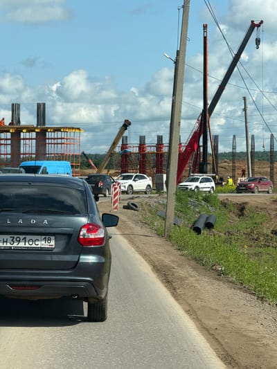 The image depicts a construction site on a sunny day with a cloudy sky. In the foreground, the rear of a dark gray Lada sedan is visible, with its license plate reading "H391OC 18 RUS". The car is on a paved road, with a dirt and grass shoulder to its right.

The construction site features several large concrete pillars under construction, with rebar exposed at their tops. Orange scaffolding is erected around some of these pillars. Two large cranes are present, one red and one yellow, actively involved in the construction process. Several workers in orange vests and hard hats are visible on the site. Other vehicles, including a blue van, a black sedan, and two white SUVs, are parked or driving on a road within the construction area. In the background, a wooded area is visible. The scene suggests ongoing infrastructure development. The location is identified as Kabanikha, Russia.