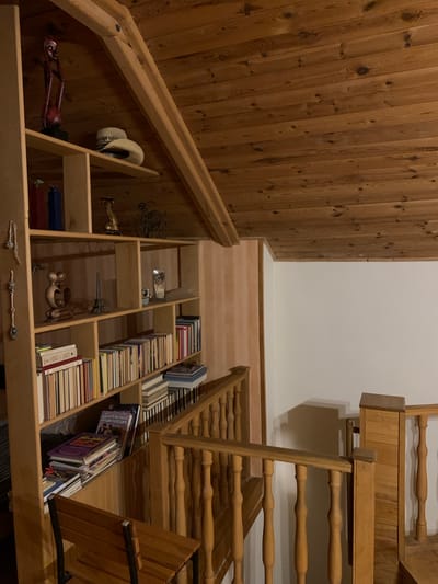 This image depicts an interior view of a room with a wooden ceiling and shelves filled with books and decorative items. The scene is likely a home or a small library. On the top shelf, a wooden figurine is displayed along with a white hat. Below this, various books are organized on different shelves. Some decorative objects, such as a miniature Eiffel Tower and a vintage bicycle figurine, are also visible. A wooden staircase with railings is present in the foreground, suggesting this might be an upper level or loft. The overall ambiance is warm and cozy due to the extensive use of wood. There are no people present in the image.