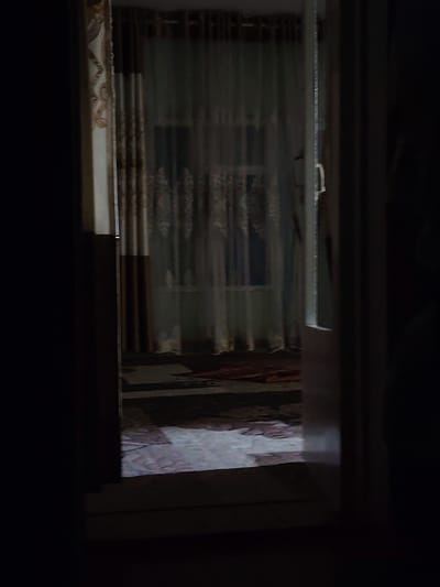 The image, captured in Akaltyn, Uzbekistan, depicts an interior scene viewed through an open doorway. The foreground is dominated by darkness, with the right side of the frame showing the edge of a wall or doorframe. The interior space beyond is dimly lit, with sheer curtains obscuring a window. The curtains have a patterned design, appearing to be a light color with darker motifs. A rug is visible on the floor, partially illuminated, showing a pattern of brown and lighter tones. The overall impression is of a residential interior, possibly a living room or bedroom, at night or in low light conditions. No people are visible in the scene.