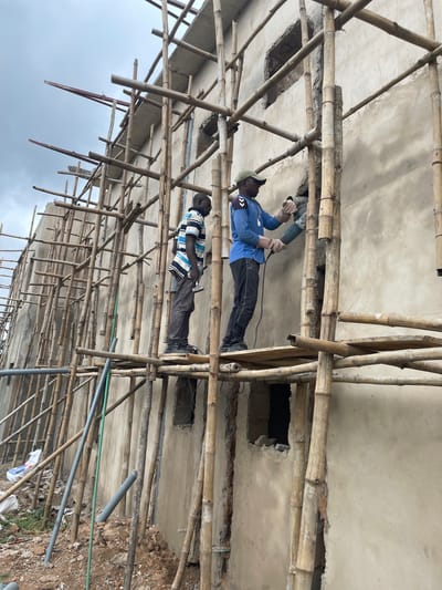 This is a photograph taken in Okinano, Nigeria, capturing a construction scene. Two men are present, working on the exterior of a building that is partially covered in scaffolding made of bamboo poles. The building's walls appear to be made of concrete and are in a raw, unfinished state, with visible gaps or openings.
The man on the right is wearing a blue long-sleeved shirt, black trousers, a green cap, and gloves. He is actively using a grinding tool, which is a power tool with a spinning disc, to work on the wall. He is standing on a wooden platform attached to the bamboo scaffolding.
The man on the left is wearing a striped shirt (blue and white) and dark trousers. He is also standing on the bamboo scaffolding, and he is holding a tool, possibly a hammer or chisel, and appears to be engaged in demolition or preparation work on the wall.
The environment is a construction site with loose rocks and debris on the ground. Several pipes, one green and one gray, are visible near the base of the building, suggesting plumbing work is also in progress. The sky is overcast, indicating a cloudy day. The overall scene depicts active manual labor in the construction of a building in Nigeria.