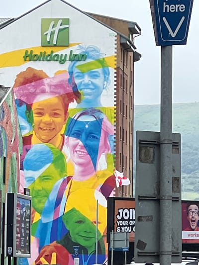 This image captures a vibrant mural painted on the side of a building in Belfast City District, United Kingdom. The mural features a collage of diverse faces rendered in bold, colorful strokes, evoking a sense of community and inclusivity. Prominently displayed on the building is the "Holiday Inn" logo, indicating the hotel's presence. In the foreground, a metal pole supports a blue sign with white lettering that reads "here" and points downwards with a white arrow, likely a directional sign for the hotel. The background shows a slightly out-of-focus view of rolling green hills under an overcast sky. To the right of the mural, another advertisement with the word "JOIN" is visible.