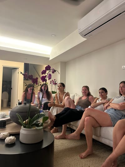 The image shows six women sitting on a white couch in a living room, located at Latitude 9.658, Longitude -84.673 in Playa Herradura, Costa Rica. The women are all dressed casually and appear to be engaged in conversation. Some women have their arms wrapped around each other, while others are sitting with their arms crossed. The room is brightly lit and decorated with a large potted plant with purple orchids. There is a white air conditioner mounted on the wall in the upper right corner of the image. One woman is wearing a white tank top and a silver necklace. The other woman is wearing a white tank top and black pants. The couch is covered in a light brown throw blanket. The room has white walls and a beige floor. There is a black coffee table in the center of the room with a plant on it. In the background, a doorway leads to a hallway with a toilet visible to the left.