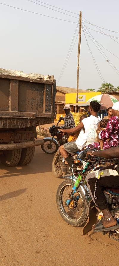 This street-level photograph captures a bustling scene in Ketu Adieowe, Nigeria, featuring vehicles and people on a dusty road under a hazy sky.

In the left foreground and midground, a large, dark-colored dump truck is prominent, its side and rear heavily caked with mud or dirt. The truck's robust tires are visible at the bottom left. Adjacent to the truck, several motorcycles are navigating the crowded space. One man in a distinctive spotted long-sleeved shirt sits on a motorcycle, looking towards the right.

Further to the right, other individuals are on or around motorcycles. A man in a white t-shirt and grey shorts sits on a bike, and behind him, a woman in a vibrant purple and white patterned dress holds a child, with another woman partially visible. The closest motorcycle on the right shows a person's foot wearing a black sandal and an anklet with small cross-like charms.

The road surface appears reddish-brown and unpaved or heavily worn, indicating a dusty environment. Shadows cast on the ground suggest sunlight, possibly from a hazy midday or late afternoon.

In the background, various buildings line the street. A notable yellow building features a clear "MTN" logo and the text "Internet Point & Registration Centre" below it, indicating a commercial establishment. A tall, wooden utility pole stands on the right side, supporting numerous tangled and exposed electrical wires, a common sight in the region. The sky is a light, uniform greyish-blue, consistent with a dusty or hazy atmosphere. The scene conveys a sense of vibrant, everyday activity and transportation in a busy local setting.