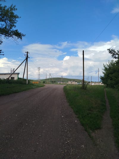 The image depicts a rural scene on a partly cloudy day, likely in or around the village of Ziyanchurino, Russia, based on the provided context. A dirt road curves from the foreground into the distance, flanked by green grass and vegetation on either side. Several utility poles with wires are visible along the roadside, one with a satellite dish attached to a building on the left. In the mid-ground, there are several low-rise buildings, suggesting a small settlement or industrial area. A tall cellular tower stands prominently in the middle distance, and rolling, green hills form the backdrop under a bright blue sky with scattered white clouds. The overall impression is one of a quiet, somewhat isolated rural landscape with signs of modern infrastructure. There are no people or animals visible in the image.