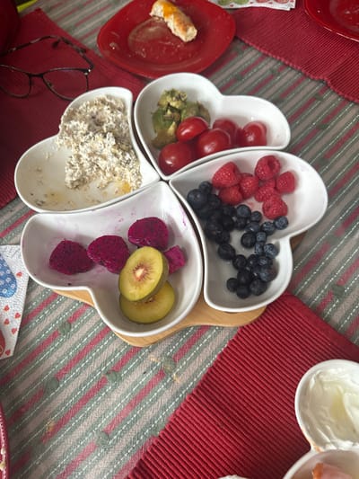 The scene depicts a tabletop setting, likely indoors, within the city of Matveevskoe, Russia. The primary subject is a four-section, white ceramic serving dish, shaped like connected hearts, positioned centrally on a light-colored wooden or bamboo base.
Each section of the ceramic dish contains different food items:
* The top-left section holds a partially consumed, creamy, beige-colored mixture with dark specks, consistent with oatmeal or a similar cereal.
* The top-right section contains green, cut avocado pieces and several whole, red cherry tomatoes.
* The bottom-right section is filled with fresh blueberries and raspberries.
* The bottom-left section displays slices of red dragon fruit and two golden kiwi halves.
Surrounding the central serving dish, other objects are present on the table:
* In the upper-left, a pair of black-framed eyeglasses rests on a red, ridged placemat.
* Adjacent to the eyeglasses, a small red square plate contains a partially consumed bread item with an orange topping and some liquid residue.
* A white napkin with a blue and red heart pattern is partially visible on the lower-left.
* In the lower-right corner, a portion of another white bowl or dish containing a white, creamy substance is visible.
The table is covered by a striped tablecloth featuring alternating light green/grey and thin red/white stripes, with small, raised grey textural elements. A red, ridged placemat is also visible on the lower-right side of the frame. No individuals are visible, and no active interactions or actions are depicted. The arrangement suggests a meal or snack is in progress or has recently been consumed.