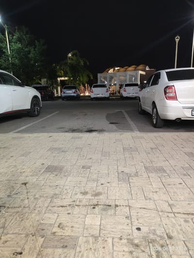 The image depicts an outdoor parking area at night, with several vehicles parked. In the foreground, the ground is paved with large, light-colored rectangular stones. Beyond the paved area is asphalt, marking parking spots. Several cars are visible, including a white SUV on the left, a light blue sedan in the middle, and two white sedans further back. A building with ornate white latticework and a curved roof is visible in the background, illuminated by interior lights. Trees are present on the left side of the image, and streetlights cast illumination. The scene appears to be near a restaurant or event venue, potentially located in the city of Turkistan, Kazakhstan, given the time stamp in the lower right corner: "2026 06 17 21:16". No people are clearly visible.