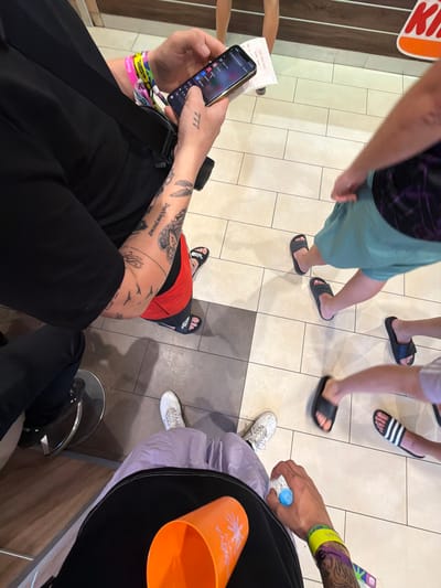 The image captures a scene with multiple individuals in an indoor setting. The flooring consists of light-colored square tiles. Several people are wearing casual footwear like sandals and slides. A person is holding a smartphone and has visible wristbands and tattoos on their arm. Another person is holding a bottle of water and a cup and is also wearing wristbands and has tattoos. In the lower right corner, a metal stool is visible. The presence of a receipt indicates a commercial establishment. The image was captured indoors in Punta Umbría, Spain.