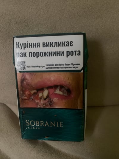 This image shows a close-up of a pack of Sobranie cigarettes. The pack features a graphic warning image of diseased oral tissue and text in Ukrainian that translates to "Smoking causes mouth cancer." Below the warning, the brand name "SOBRANIE" and "LONDON" are visible in gold lettering on a dark teal background. The pack appears to be placed on a textured surface, possibly a couch or carpet, in an indoor setting. There is no visible indication of the location being Skarydzew, Poland.