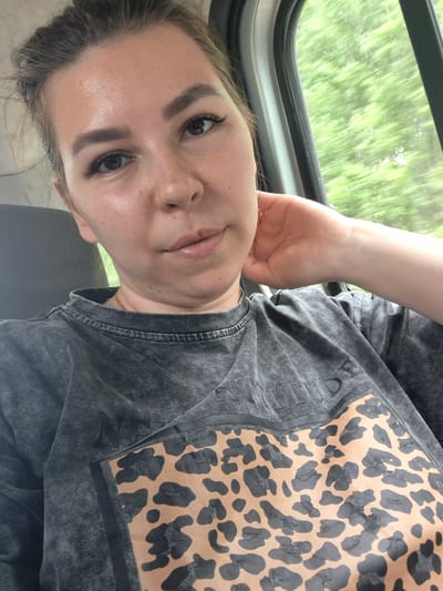 A young woman is seated inside a vehicle. She is wearing a grey t-shirt with a leopard print design on the left side. She is holding her face with her right hand, resting her elbow on the windowsill. Through the window, blurred green trees are visible.
