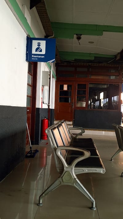 The image depicts an interior space, likely a train station or similar transit hub, in Karawang, Indonesia. The foreground features rows of metal and wood waiting benches affixed to a tiled wall. In the midground, a blue sign with an icon and the words "Keamanan Security" (security in Indonesian) sits on a white surface. Behind this is a brown wooden structure resembling a partitioned door or window frame. A red fire extinguisher is positioned near the benches. In the background, there is a glimpse of an outdoor area through the wooden structure, indicating a potential entrance or exit. A mop is visible, suggesting recent or ongoing cleaning activity. The overall scene is one of a functional public space.