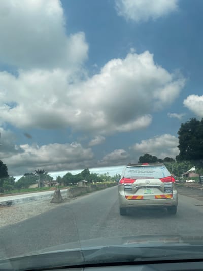 This media file shows a view from inside a vehicle, looking out at a road and the sky. The primary subject is a gold-colored minivan driving on a paved road. The minivan has a large decal on its back with text that includes company information and contact numbers. Text visible on the van reads "JOKIMNA MOTORS," followed by a list of cities like "ABUJA," "ENUGU," "PORT-HARCOURT," "BENIN," "UYU," "LAGOS," "WARRI," "BAYELSA," "CALABAR," "JOS," "KANO," and "KADUNA." It also includes a website address "www.jokimnamotors.com" and phone numbers "08100079208, 09022658295." There are also smaller logos or text that say "DUNAMIS" on the rear of the van. The road appears to be in a rural or semi-urban area, with trees and some simple buildings visible on the side. The sky is a prominent feature, filled with dramatic clouds against a blue sky, suggesting a dynamic weather condition, possibly before or after rain. The lighting indicates it is daytime, though the cloud cover creates a mix of light and shadow. The image seems to capture a moment of travel through Nigeria, given the city names listed on the vehicle and the provided location context.