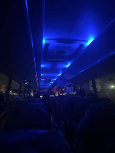 The image depicts the interior of a vehicle, likely a bus or train, viewed from a low angle and rotated clockwise, creating a vertical composition. The scene is illuminated predominantly by bright blue LED strip lights recessed along the upper section of a long wall on the left, casting a strong blue glow across the interior.
On the left side, the vehicle's interior wall is visible, featuring several square or rectangular recesses with rounded corners, each appearing to contain an unlit screen or panel. The blue light originates from strips running along the top and bottom edges of these recesses, as well as along the base of the wall.
On the right side of the image, the dark silhouettes of multiple passenger seats are visible, suggesting people are seated or have been seated there. Several indistinct figures can be seen, heavily obscured by shadow, with one person's head and shoulder partially visible in the foreground. Above the seats, small square displays or screens are mounted; one clearly shows two red symbols: a crossed-out cigarette and a crossed-out speaker, indicating "no smoking" and "no phone calls" or "silence."
The overall atmosphere is dark and modern, characteristic of nighttime travel or a vehicle designed with mood lighting. The blue lighting dominates the color palette. Given the location context of Montmagny, France, the image likely captures a moment inside a mode of public transportation in or around that area.