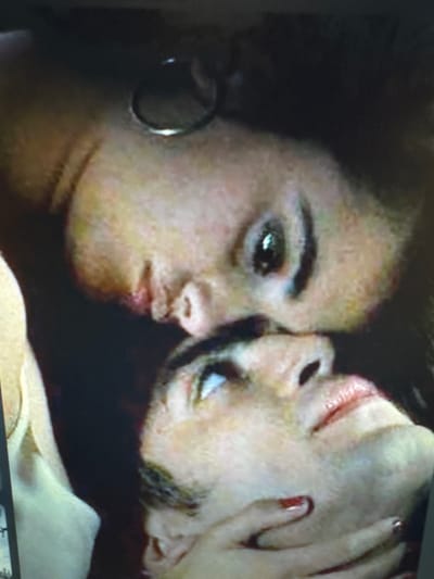 The image is a close-up, low-resolution photograph of two individuals in what appears to be an intimate moment. The focus is on their faces, with one person positioned above the other. The lighting is dim, and the overall quality of the image is poor, suggesting it may be a still from a video or a poorly taken photograph. There is no visible indication of the location being Protasovo, Russia, or any other specific city. The individuals' faces are partially obscured and blurry, making it difficult to ascertain specific details about their appearance or expressions. One individual has dark hair and is wearing what appears to be an earring. The other individual's hand is visible near their chin, with painted fingernails. The background is dark and indistinct.