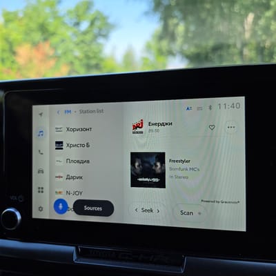 The image shows a car's infotainment system displaying the FM radio station list. The screen shows several radio stations with their names and frequencies, including "Енерджи" at 89.50. The time displayed is 11:40. The system indicates it is connected to FM and shows a station list. There is also a song playing, identified as "Freestyler" by "Bomfunk MC's". The bottom left of the screen shows volume controls and a settings icon. The car appears to be in an outdoor environment, as indicated by the blurry green and orange foliage visible through the windshield. The location of Ravnogor, Bulgaria is not discernible from the image.