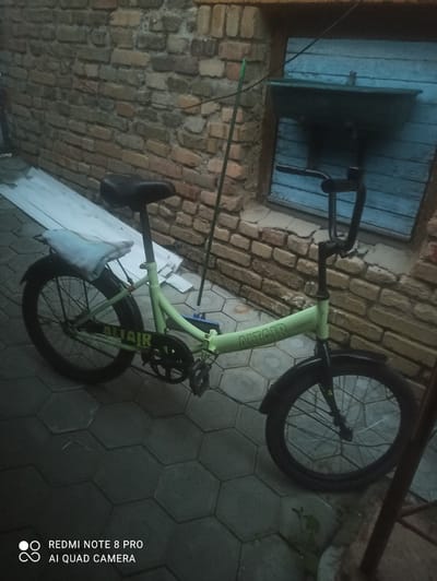 The image depicts a light green children's bicycle with the brand name "ALTAIR" visible on the chain guard and frame. The bicycle is parked on a patterned stone-tiled surface next to a brick wall. To the right of the bicycle, a blue wooden structure is partially visible. A white sheet is draped over the rear of the bicycle seat. In the bottom left corner, there is text that reads "REDMI NOTE 8 PRO AI QUAD CAMERA", indicating the device used to capture the image. The scene appears to be an outdoor area, possibly a courtyard or alley, in a city. The specific location within the city of Irbit, Russia, cannot be determined from the image alone.