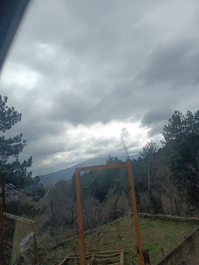 The image captures an elevated, outdoor landscape view identified as being from or near the city of Yavrovo, Bulgaria.

The upper half of the frame is dominated by a heavily overcast sky, filled with dense, grey stratus clouds. Brighter patches within the cloud cover suggest areas of diffuse light penetration. A dark, blurry, diagonal obstruction is visible in the extreme upper left corner of the image, extending downwards.

In the foreground, a prominent, vertically oriented, rectangular structure constructed from rusty, dark orange-brown metal stands centrally. This structure appears empty, framing the landscape beyond it. The immediate ground around the metal frame consists of dry, sparse green grass, exposed earth, and scattered dry brush. To the left and right of the frame, and extending into the midground, are slopes densely covered with a mixed forest of evergreen coniferous trees (primarily pines) and numerous bare deciduous trees, suggesting a cooler season.

A significant reflective glare or lens flare obscures the majority of the lower left quadrant of the image, rendering underlying details indistinct. Faint outlines consistent with a window pane or another reflective surface are minimally discernible beneath this glare. The lower right foreground reveals the edge of a concrete or stone structure, possibly a wall or foundation.

In the background, multiple layers of mountains or hills recede into a light atmospheric haze. No individuals, animals, or explicit actions are depicted within the scene.