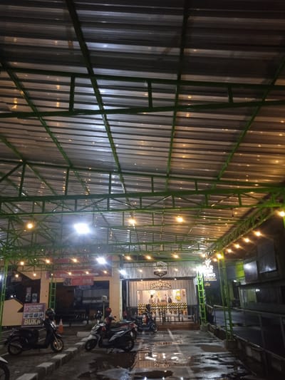 This nighttime image captures an outdoor commercial area, likely a food court or a strip of small businesses, located in Kecamatan Kartasura, Indonesia. The scene is dominated by a large, open-air canopy featuring a green metal framework supporting a corrugated metallic roof. Numerous warm string lights are strung across this structure, illuminating the space below.
The ground is paved and appears wet, possibly from recent rain, with white painted lines indicating parking spots. Several motorbikes of various makes are parked in the foreground, suggesting customer or staff presence.
On the left side of the frame, a food stall or restaurant is visible, marked by signs and banners. A prominent sign partially reads "Sate Kambing & Tengkleng" (Goat Satay & Tengkleng), and another sign below it clearly states "BUKA" (Open). A large red banner with white text is displayed above this establishment.
To the right, a modern barbershop named "BARBER KING" is clearly visible, with its distinctive logo and white-and-black striped facade. Large glass windows allow a view into its interior. The artificial lighting against the dark backdrop confirms the time of day as night. The overall atmosphere suggests a lively evening commercial setting.