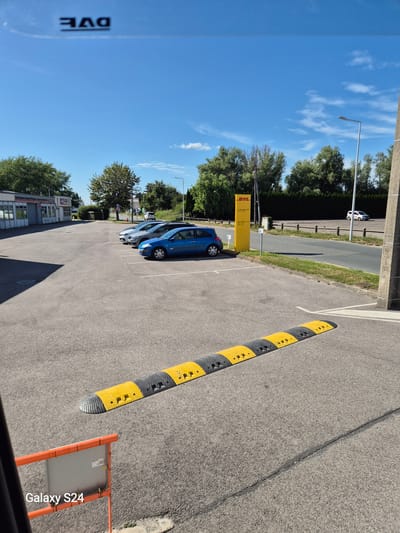 This is a landscape orientation photograph taken outdoors on a sunny day, likely in the afternoon given the shadows. The image is dominated by a grey asphalt parking lot with a prominent yellow and black speed bump in the foreground, running diagonally across the frame. To the left of the speed bump, several parked cars are visible, including a blue hatchback. In the background, there is a row of trees and a DHL sign with yellow and red branding. Further left, a street lamp stands tall against a clear blue sky with scattered white clouds. The image appears to be taken from a slightly elevated perspective. Some text is visible: "DVE" on the left side of the frame, and "Galaxy S24" in the bottom right corner, suggesting the photo was taken with a Samsung Galaxy S24 phone. The overall scene depicts a typical parking area, possibly near a business or distribution center, given the DHL sign. There are no people visible in the frame, and the atmosphere is calm and bright.