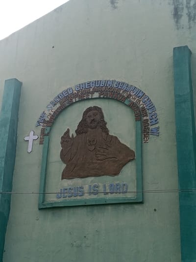 The image shows a religious building or church exterior in Jibowu, Nigeria. A green concrete wall features a mounted, textured depiction of Jesus Christ in brown. Above the image, arched text reads "THE SACRED CHERUBIM/SERAPHIM CHURCH W/ MEMBER OF C UNIFICATION BINUKONU PARISH MOUNT GOSHEN STREET PANADA AGEGE." Below Jesus, the words "JESUS IS LORD" are inscribed. A white cross is located on the left side of the image. The building appears to be weathered, with some discoloration and imperfections on the wall. Two greenish pillars stand on either side of the image and the building wall.