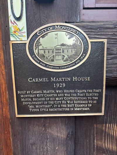 A rectangular bronze-colored historical plaque is mounted vertically on a dark brown wooden surface with prominent grain, likely a door or wall, in the city of Monterey, United States of America. The plaque features a raised border and text in a lighter, polished bronze tone, contrasted with a darker, textured background.

The top portion of the plaque is an oval section framed with a raised border. Inside this oval is an engraved image depicting a two-story building with a central entrance and a flagpole flying a flag, surrounded by trees. Above this image, in an arch, are the words "CITY OF MONTEREY". Below the image, following the curve of the oval, is the text "Colton Hall, California Constitution Hill 1849".

Below the oval section, the main body of the plaque contains the following raised text:
"CARMEL MARTIN HOUSE"
"1929"
"BUILT BY CARMEL MARTIN, WHO HELPED CREATE THE FIRST MONTEREY CITY CHARTER AND WAS THE FIRST ELECTED MAYOR. BECAUSE OF HIS MANY CONTRIBUTIONS TO THE DEVELOPMENT OF THE CITY HE WAS REFERRED TO AS "MR. MONTEREY". IT IS THE BEST EXAMPLE OF TUDOR STYLE ARCHITECTURE IN MONTEREY."

To the upper left of the plaque, a portion of a decorative ceramic tile is visible, featuring a floral design with yellow, red, and green elements on a pale background. The scene is illuminated from the right, casting faint shadows.