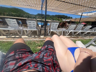 The image shows a sunny day at a poolside resort. In the foreground, a couple is relaxing on lounge chairs. The person on the left is wearing camouflage-patterned swim trunks, and the person on the right is wearing a blue bikini bottom. In the background, there are rows of white lounge chairs under a white sunshade. Beyond the chairs, a blue swimming pool is visible, and behind that, lush green hills dotted with houses. The location is identified as Delchevo, North Macedonia.