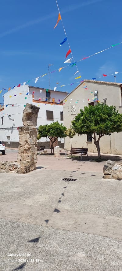 The image shows a street scene in Canals, Spain. The sky is clear blue. A string of colorful triangular pennants are strung across the street. The buildings are primarily white with some beige. There are two green trees on the right side of the image, next to a beige building with two benches. A stone pillar stands in the foreground. The ground is paved with gray concrete. The shadows of the flags can be seen on the ground. A white car is partially visible at the bottom. A camera watermark indicates the date: June 1, 2026.