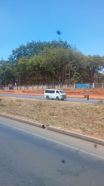 A white van drives along a newly paved road under a clear blue sky. The road is bordered by a grassy patch and a lower street. A concrete fence and dense green trees line the background, partially obscuring buildings and utility poles. There are dark spots appearing in the image, especially in the sky and the road.