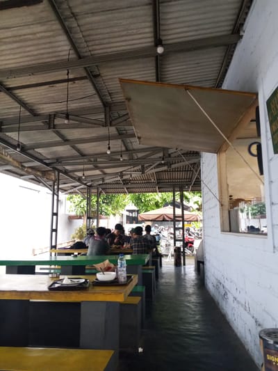 The image depicts an outdoor dining area at a casual eatery, likely a cafe or restaurant, located in Kelurahan Merjosari, Indonesia. The scene is viewed from a low angle, looking towards the right. The roof is constructed from corrugated metal sheets supported by a metal frame structure. Several simple pendant light bulbs hang from the ceiling.

In the foreground, there are several picnic-style tables with yellow and green colored seats and tabletops. One table in the center has a plastic tray with a white bowl, a white napkin, and a bottle of water. Another tray on the same table holds a small plate with some white food and an ashtray.

Further back, there are more tables where several individuals are seated, engaged in eating and socializing. The patrons are dressed casually. Some are looking at their phones, while others are interacting with each other. Beyond the seating area, there are trees and foliage, suggesting an outdoor or semi-outdoor setting. In the background, a row of parked motorcycles indicates a common mode of transportation in the area.

To the right of the image, a white brick wall runs along the side of the establishment. A window with a blue clock showing approximately 9:10 is visible in the wall. A sign with green lettering and the word "SIGN" at the bottom is partially visible on the wall to the far right. The floor is wet and reflective, suggesting recent rain or cleaning.