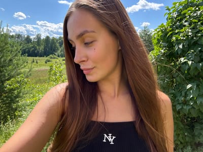 A young woman with long, brown hair is photographed outdoors in a rural setting, likely on a sunny day. She is wearing a black tank top with a white "NY" logo. Her head is tilted down and to her right, with her eyes closed. The background consists of lush green trees, fields, and a bright blue sky with scattered white clouds. The location is described as the city of Sholokhovo, Russia.