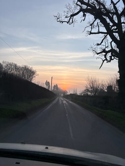 A forensic examination of the image reveals a rural road scene in Ruyton-XI-Towns, United Kingdom, at either sunrise or sunset. The perspective is from inside a vehicle, with a dark dashboard visible across the bottom of the frame. The asphalt road surface is wet, reflecting the sky's light, and features a faded white dashed center line extending into the distance.
The sky occupies the upper portion, displaying a gradient from pale blue and light grey at the top to vibrant orange and yellow hues at the horizon. A bright, circular orange sun is visible low on the horizon, partially obscured by distant trees and atmospheric haze, indicating the low angle of the sun.
Along the left side of the road, a dark, dense hedgerow or line of bare trees forms a continuous barrier. Behind this, additional bare deciduous trees are silhouetted against the lighter sky. A utility pole with a single overhead wire extends from the left foreground upwards.
On the right side of the road, a low stone wall covered in dark green vegetation, possibly ivy, runs parallel to the roadway. Behind this wall, several bare deciduous trees and the dark silhouette of a building or structure are visible. Dominating the upper right quadrant is a large, dark, gnarled deciduous tree with numerous bare branches extending into the sky.
No human figures, animals, or other active vehicles are visible in the scene. The wet road surface and atmospheric haze contribute to a slightly muted and reflective environment.