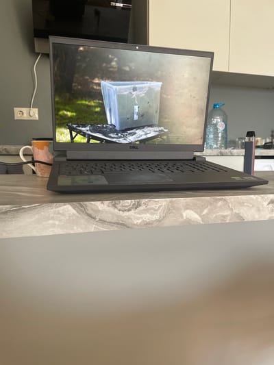 The image depicts a modern kitchen counter with a Dell laptop open. The laptop screen displays an image of a clear plastic container with plants and soil, placed on a black folding table outdoors. To the left of the laptop, a mug with an orange and pink marble pattern is visible. To the right of the laptop, a large plastic bottle of water and a vape pen are on the counter. In the background, a wall-mounted electrical outlet and a portion of a dark television screen can be seen. The kitchen features light-colored cabinets and a countertop with a grey and white marble pattern. The scene is indoors, in a kitchen environment. The city of Obninsk, Russia, is not explicitly indicated in the visual information provided in the image.