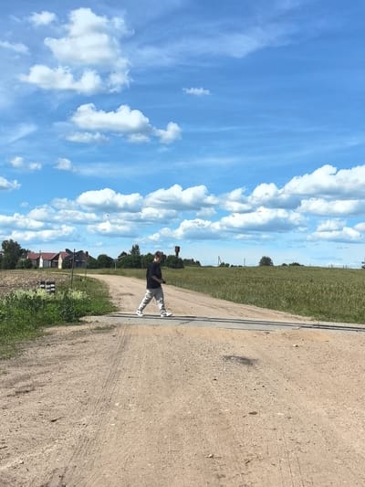 A lone individual is walking across a dirt road that has a set of railroad tracks running across it. The person is wearing a black t-shirt and light-colored, patterned pants, and white sneakers. They appear to be holding a small, dark object, possibly a camera, in their right hand. The scene is outdoors, set in a rural environment. In the background, there are several houses with red roofs, surrounded by green trees and fields. The sky is a vibrant blue, dotted with fluffy white clouds, suggesting fair weather and daytime. The ground is a mixture of dirt and gravel, with some tire tracks visible. The overall impression is one of a peaceful, sunny day in the Belarusian countryside. There is no visible text in the image.