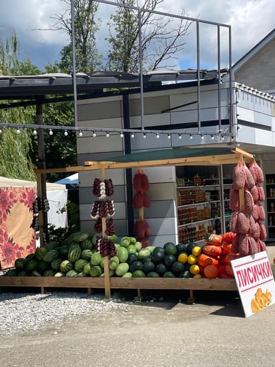 The image depicts an outdoor market stall in Gorskoye, Russia. The stall is laden with a variety of produce, including watermelons, pumpkins, and what appear to be dried sausages hanging from the stall. There are also strings of onions or similar bulb vegetables hanging. Behind the produce, shelves inside the stall are stocked with jars, likely containing preserves or honey. A sign in the foreground with Cyrillic lettering and an image of dried mushrooms indicates that the mushrooms, "лисички" (lisichki), are for sale. The stall is set up on a gravel surface, and the background shows trees and a building. The sky is partly cloudy. No people are visible in the image.