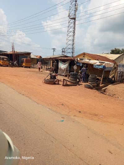 The image shows a dirt road in front of a tire repair shop, located in the town of Aramuko in the state of Ogun, Nigeria. The shop is on the right side of the road and is characterized by an open-sided structure with a blue tarp canopy. A tall metal tower with a large amount of power lines is visible in the background. There are two figures standing near the tire shop, and a third figure walks toward the road in the distance. A motor is visible on the ground, likely used to inflate tires. Stacks of tires and other tools are stored along the side of the shop. There are three-wheeled vehicles parked in front of the shop. The ground is dirt and dusty, with some grass growing on the edges. The sky is blue with white clouds.