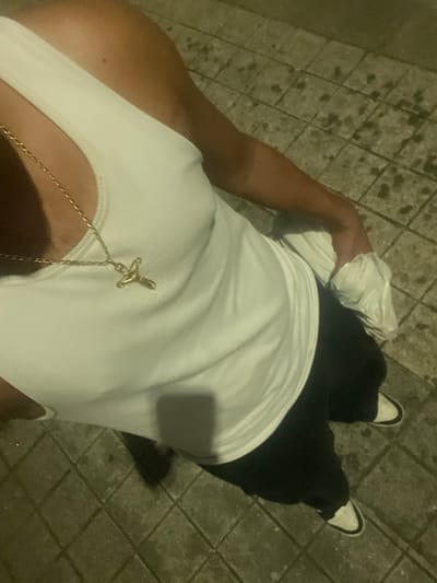 A high-angle, downward perspective captures the upper body and legs of an individual standing on a tiled pavement in Pedrouços, Portugal. The subject wears a white ribbed tank top, a gold-colored chain with a crucifix pendant, black trousers, and white sneakers with black accents. The right hand holds a crinkled white plastic bag at hip level. The pavement consists of square, light-colored concrete or stone tiles, exhibiting multiple dark, damp-appearing stains. Shadows indicate low or artificial light sources.