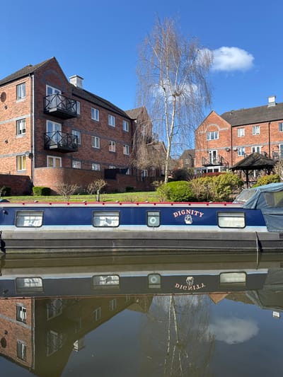 A sunlit scene depicts a tranquil canal environment, likely within the Wyre Forest district of the United Kingdom. A dark blue and black narrowboat named "DIGNITY," featuring a white graphic of a traditional canal boat, floats on calm, reflective water. Its reflection, along with the surrounding architecture and a bare-branched tree, is visible in the water.
On the canal bank, a series of red-brick residential buildings, appearing to be multi-story apartments or townhouses with dark roofs and white-framed windows, line the waterway. The building on the left features two prominent balconies with black railings. A tall, slender deciduous tree with bare branches stands between the buildings. Further right, a hexagonal wooden gazebo with a dark roof is visible amidst green foliage. The ground slopes gently upwards from the canal, covered with green grass and shrubs. The sky is clear blue with a single white cloud present on the right side. No individuals or active interactions are visible in the scene.