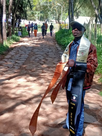 An outdoor scene in Waseges ward, Kenya, depicts a young male in the foreground, positioned on the right, looking directly at the camera. He wears a beige baseball cap, a blue denim shirt, a reddish-brown patterned jacket, and blue jeans. He holds multiple academic sashes: a cream-colored sash over his left shoulder inscribed "LAIKIPIA UNIVERSITY" and "CLASS GRAD," a black sash on his right side marked "LAIKIPIA UNIVERSITY" and "CLASS OF 2025," and two long, reddish-brown sashes hanging downwards, one displaying "CLASS of 2025" and "Junior Graduate."
Behind the primary subject, a group of approximately six to eight individuals walks away from the viewer along a winding path. The path is constructed from irregular flagstones or cobbles embedded in dirt. These individuals are casually dressed, with one person notably wearing orange trousers. The environment is characterized by lush green vegetation, including tall trees and bushes, bordering both sides of the path. Papers or posters are affixed to a tree trunk on the left. In the mid-ground, a white utility pole stands on the right side of the path. Distant structures with light-colored walls and dark roofs are partially visible through the trees in the background. The scene is brightly lit by sunlight, casting prominent shadows on the ground.
