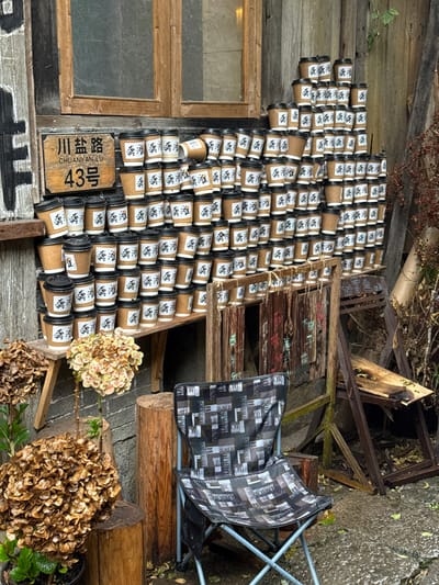 The image captures an outdoor scene at 43 Chuanyan Road (川盐路), Gongtan, China, against a weathered wooden building facade featuring a window. Hundreds of disposable coffee cups, predominantly brown kraft paper with white labels displaying black Chinese characters (likely "龚滩"), are meticulously stacked in a multi-tiered display on a wooden shelf and directly against the wall. Most cups are fitted with black plastic lids.

To the left of the main coffee cup display, a wooden street sign is affixed to the wall, reading "川盐路 CHUANYAN LU 43号". A tall, dark wooden sign with white vertical text is partially visible on the far left. Below the extensive cup arrangement, a smaller wooden rail supports several distressed wooden planks with faded painted characters; one plank features red characters indicating "始营业中" (Now Open).

In the foreground, a blue-framed foldable camping chair with a black and grey patterned fabric seat and backrest is positioned on damp, stony ground. Several wooden stumps are scattered, with one supporting a potted plant containing dried hydrangea-like flowers. Additional dried foliage is visible on a wooden shelf to the left and along the right edge of the frame, where a derelict wooden structure with broken planks is also present. No individuals are visible in the scene.