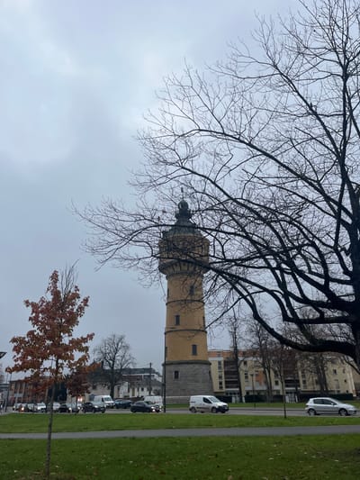 A tall tower, partially obscured by bare tree branches, dominates the landscape under a cloudy sky. The tower transitions from light yellow brick to gray stone. Cars and buildings line a street beside a narrow green strip of grass. An additional tree with brown leaves stands near the street, partially framing the lower right. The overall scene appears to be in an urban or suburban setting.