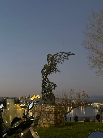 This evening image captures a serene waterfront scene in Chalkida, Greece, under a twilight sky. The central subject is a large, intricate metal sculpture, possibly abstract or resembling a winged or organic form, which stands on a rough stone base on the left side of the frame. Its open, skeletal structure is silhouetted against the darkening sky.
To the right of the sculpture, a grassy bank slopes down to a paved promenade running alongside a calm body of water, likely a sea or lake. A few indistinct figures of people can be seen near the water's edge, some appearing to be seated on benches, observing the view. Across the water, the distant cityscape glows with numerous artificial lights, reflecting softly on the water's surface.
The sky is a clear, deep blue-grey, indicating either dusk or early night. Bare tree branches are silhouetted against the upper left portion of the sky, while other trees with sparse leaves line the promenade. In the immediate foreground on the lower right, the handlebars and windshield of a scooter are partially visible, suggesting the photograph was taken from a parked position. No discernible text is visible in the image. The scene conveys a peaceful, contemplative mood with the interplay of natural and artificial light.