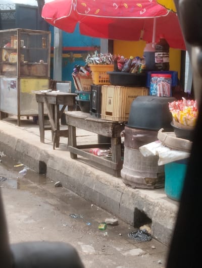 The image depicts a street scene in Oworonshoki, Nigeria, characterized by a red and yellow umbrella providing shade over a collection of merchandise. In the foreground, a wooden table with a metal structure stands on a concrete curb. To the right, another wooden table is visible, laden with various items including a large black pot, a brown pot, and several plastic containers and bags. A collection of packaged snacks or sweets in red and yellow wrappers is prominent in a black basin. Behind these items, a tall, cylindrical, patterned object, likely a large plastic container or drum, is stacked with another large black basin on top. Further back, a display cabinet with multiple shelves holds various goods. A large black plastic container with a red lid is also present. The ground is wet and littered with debris, including plastic bags and wrappers. A drain opening is visible in the concrete curb. The overall environment suggests a bustling market or street vendor area.