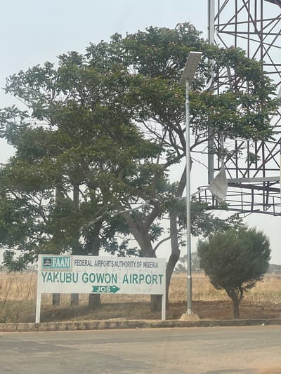 The image depicts a road sign indicating the location of Yakubu Gowon Airport in Jos, Nigeria. The sign, which is partially obscured by a large tree, also shows the logo of the Federal Airports Authority of Nigeria (FAAN). A metal lattice structure is visible behind the tree, and a small, rounded bush stands further down the road. The environment appears arid with dry grass. The image is somewhat distorted, suggesting it was taken at an angle.