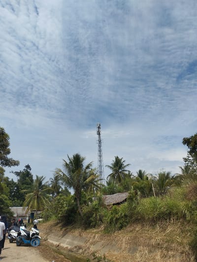 This is a wide-angle, eye-level shot taken outdoors, likely in Blangrambong, Indonesia, on a bright, cloudy day. The scene depicts a rural or semi-rural environment with a prominent cell tower in the background. In the foreground, a dirt path runs alongside a concrete ditch filled with water. Several motorcycles are parked on the path, with one white and blue scooter in the center of the frame, its kickstand down. A person in a white shirt and black pants is walking away from the camera on the left side of the path. Behind the motorcycles, a group of people are gathered, some seated on the bikes. The landscape is dominated by lush green vegetation, including numerous palm trees, and dense bushes. In the mid-ground, there is a small structure with a thatched roof. The sky is a vivid blue with wispy, white clouds stretching across it. The overall impression is a sunny, tropical day with elements of modern infrastructure amidst a natural setting.