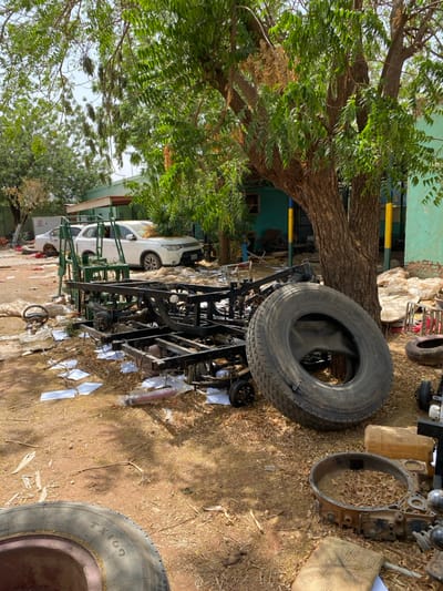 The image shows a scene in Khartoum, Sudan (latitude: 15.552, longitude: 32.57) featuring a disassembled metal trailer frame lying on the ground. The frame is partially obscured by a large tree trunk, the branches of which extend into the image. The scene is characterized by loose debris and scattered papers and documents on the ground. A white sedan is visible in the background, partially obscured by the tree's branches. The frame is partially obscured by a large tire lying on the ground next to it. The scene is situated in a residential area with a building and a grassy area in the background.
