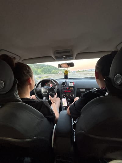The image shows the interior of a car, looking forward from the back seats. A young woman with her hair in a bun is driving, and a young man is in the passenger seat. Both are wearing black t-shirts. The car is on a highway, with green hills and trees visible outside the windshield. The sky is a light orange, indicating either sunrise or sunset. A phone is mounted on the dashboard, displaying a map application. A car air freshener is hanging from the rearview mirror. In the distance, a truck is visible on the highway.