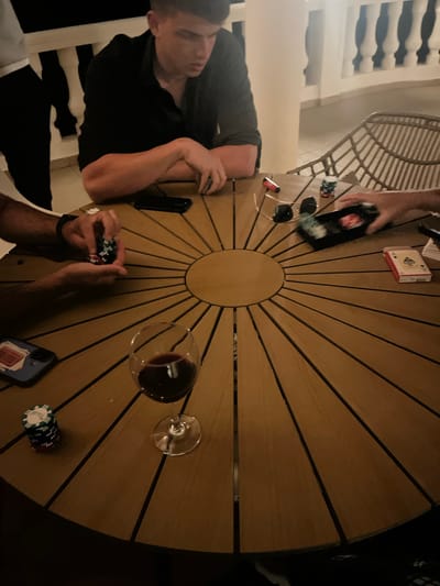 Captured from an elevated angle, the image portrays a social gathering in el Poble Nou de Benitatxell / Benitachell, Spain. A group of people are seated around a circular wooden table with a sunburst pattern, engaged in a poker game. Objects on the table include playing cards, poker chips, smartphones, a bottle of liquor, sunglasses, a lighter, and a glass of red wine. At least three individuals are partially visible: one with his arms resting on the table, and two others whose hands are visible, one of whom is holding poker chips. The surrounding environment includes a wicker chair and a white balustrade. The scene suggests an evening or nighttime setting, possibly on a patio or terrace.