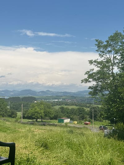 This image captures a vibrant rural landscape, likely Balabanovo, Bulgaria, under a bright blue sky with prominent white clouds. The scene is illuminated by daylight, suggesting a mid-day setting.

In the upper left corner, leafy green tree branches extend into the frame, providing natural framing. The sky, occupying the top portion, transitions from clear blue to areas with soft, voluminous white clouds, some appearing wispy. Small dark specks, possibly birds or insects, are visible against the sky.

The middle ground reveals a broad valley with a distant, undulating mountain range stretching across the horizon. Below these mountains, a small settlement or village with scattered buildings is visible. The landscape is dominated by lush green fields and dense patches of trees.

In the lower and mid-right sections, a dirt path traverses through tall green grass, dotted with small white wildflowers. Along this path, a wooden utility pole stands upright. Further along the path, several objects are visible: a small shed with a green roof, a yellow container, and a dark-colored SUV or truck parked near what appears to be a gate. A red object, possibly a small container or trailer, is also present near the path. In the immediate bottom right foreground, a portion of a dark outdoor chair or bench is visible.

The image portrays a serene natural environment with no specific human activity, conveying a sense of peace and tranquility. No visible text is present.